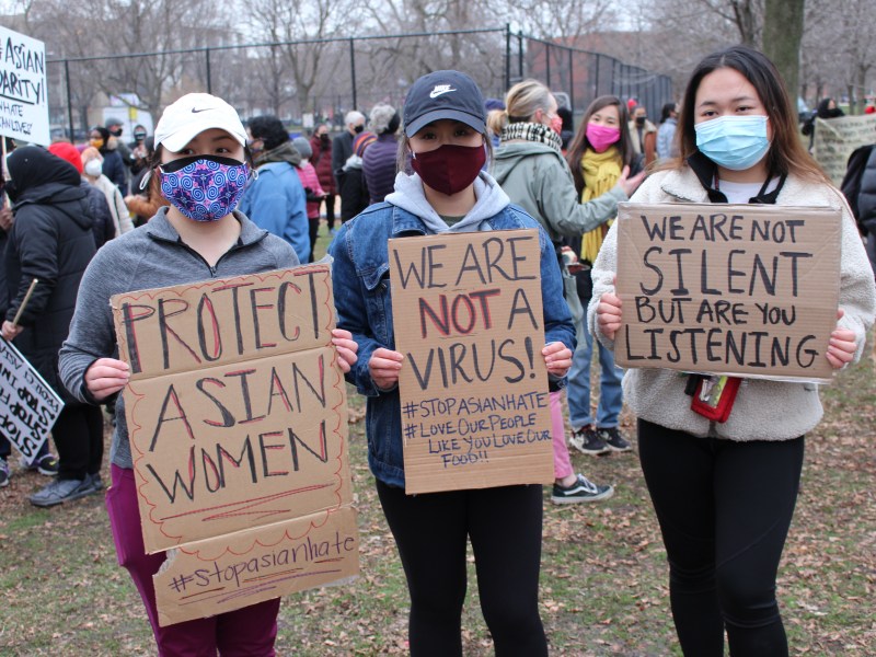 Three women hold signs at a rally against anti-Asian hate. One reads "Protect Asian Women," one reads "We are not a virus," and the third reads "We are not silent, but are you listening."