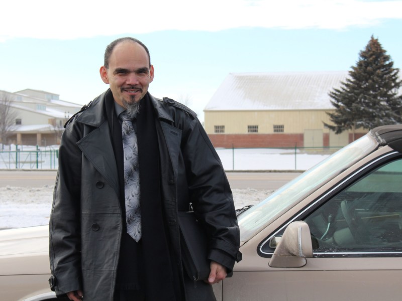Un hombre con una barba de chivo canela, vestido con saco negro y corbata, se para frente a un automóvil "lowrider" marrón.