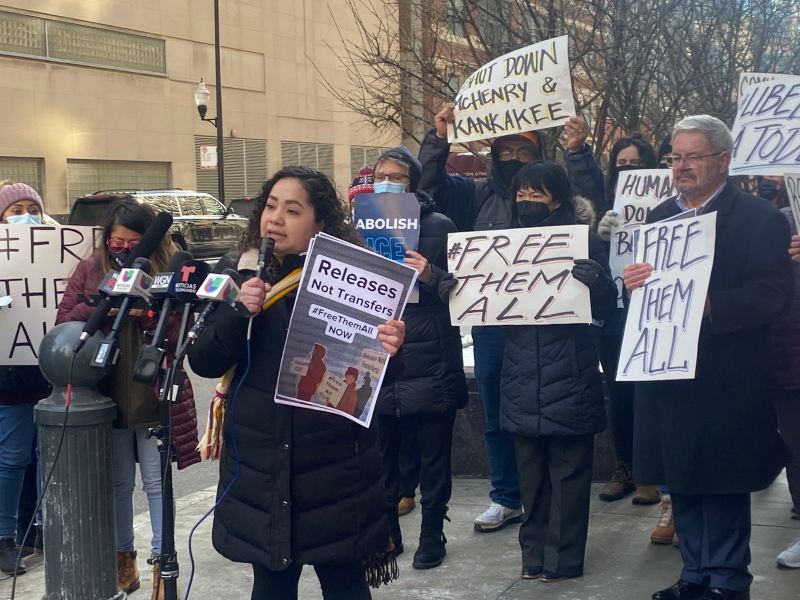 A woman stands at a microphone with people behind her holding signs that say "Free them all"