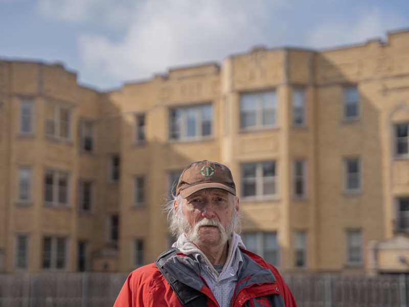 An older man with grey hair and a grey mustache stands outside in a red jacket and brown hat. There's a light brick building in the background.