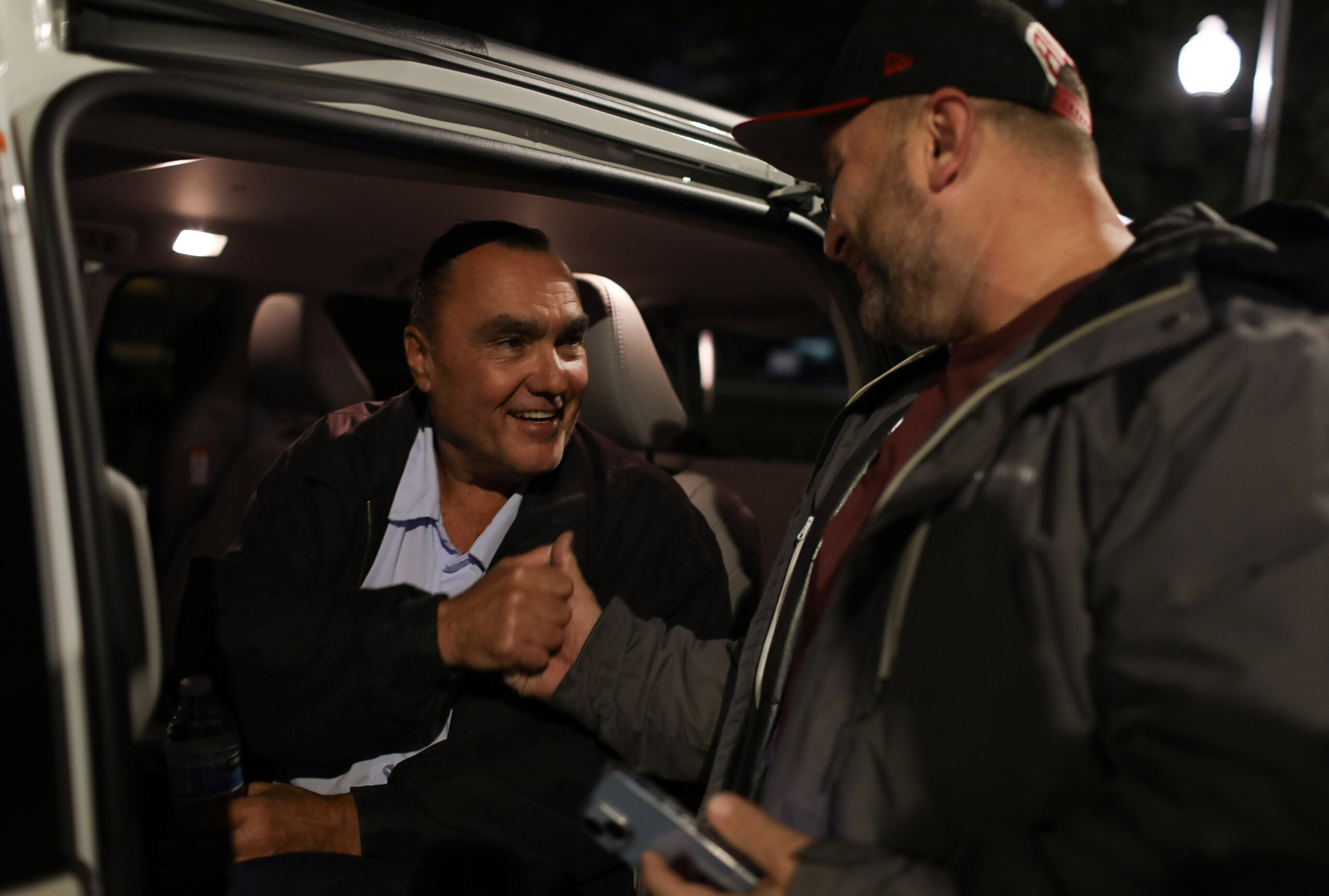 Two middle-aged men shake hands. Ronnie Carrasquillo is sitting in a car while his friend Carlos Colon stands outside.