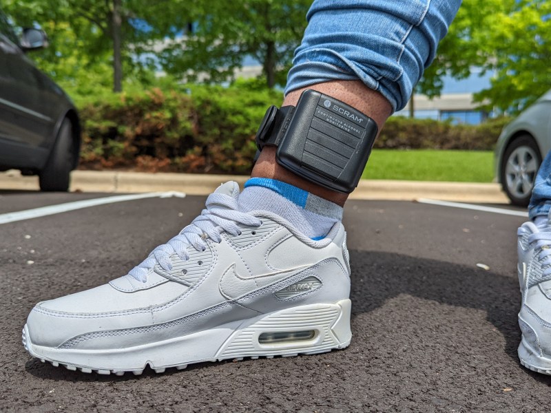 A close-up shot of a person's leg with a black square monitor strapped to their ankle. The monitor says "SCRAM continuous alcohol monitoring" on it.