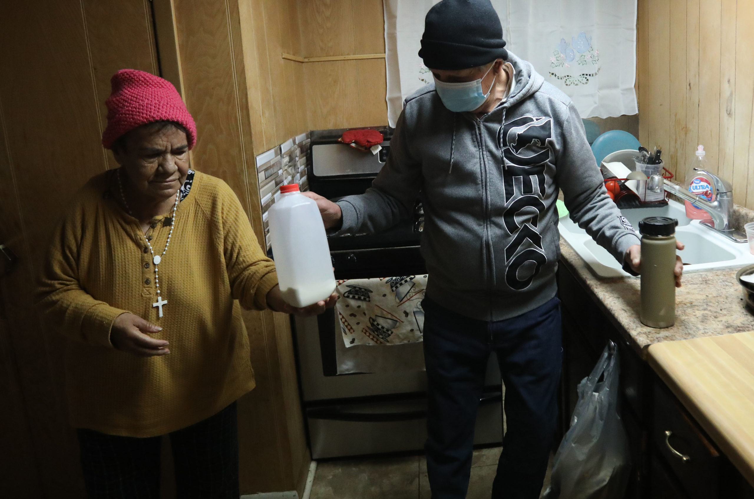 A man hands his wife a nearly-empty carton of milk while he holds a thermos in his other hand. They're both wearing winter hats and standing in a small kitchen.